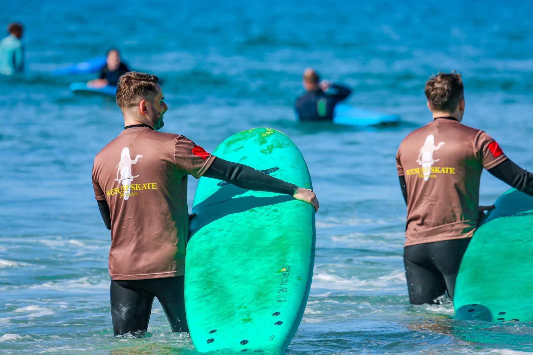 Camp surf coaches in branded rashguards in the water at Tamraght
